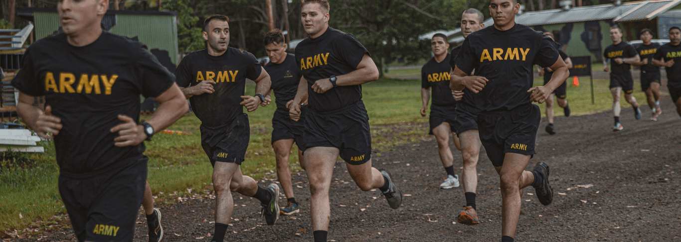 US Army soldiers in PT uniforms run the ACFT two-mile event on a dirt track surrounded by greenery.