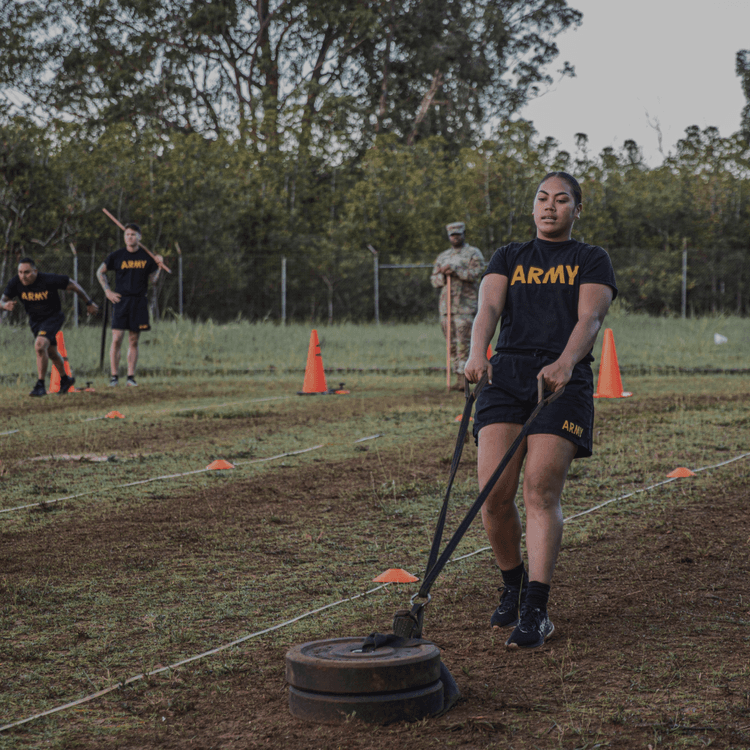 Female soldier pulls sled in Sprint-Drag-Carry ACFT event, outdoor grass field.