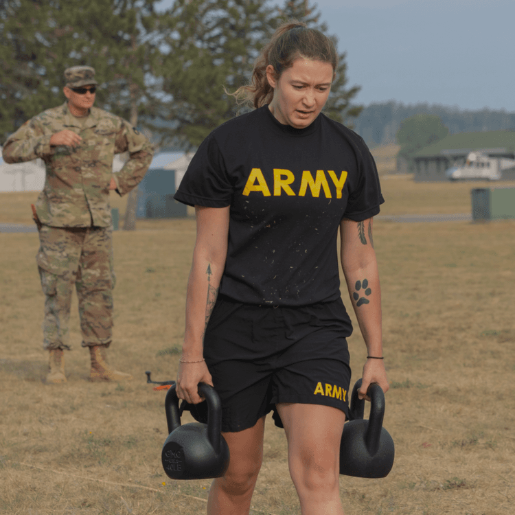 Focused female soldier carries kettlebells in ACFT event with supervisor watching.