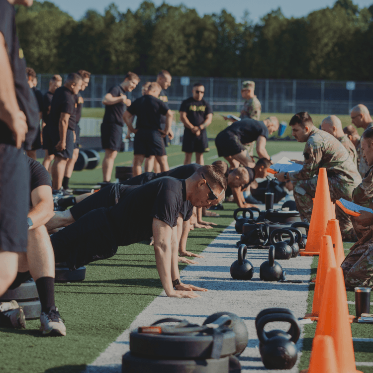 Soldier doing push-ups with others awaiting turn at ACFT on a sunny field.