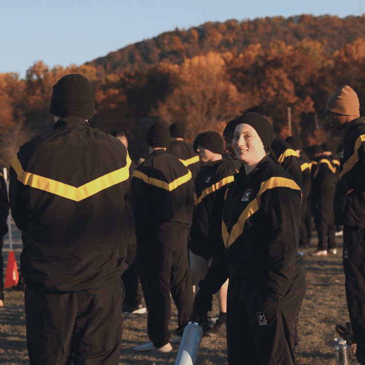 Smiling female soldier in winter PT uniform with others at dawn ACFT prep.