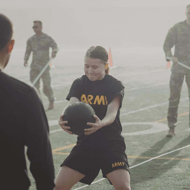 Female soldier performing medicine ball throw at ACFT in misty morning light.