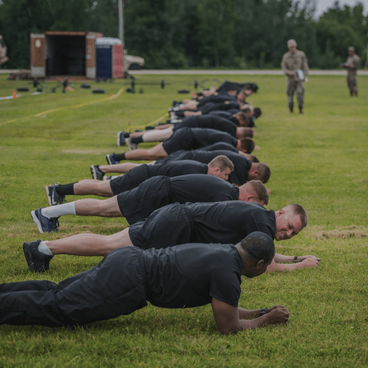 Line of soldiers doing prone planks during ACFT on a grassy field with equipment.