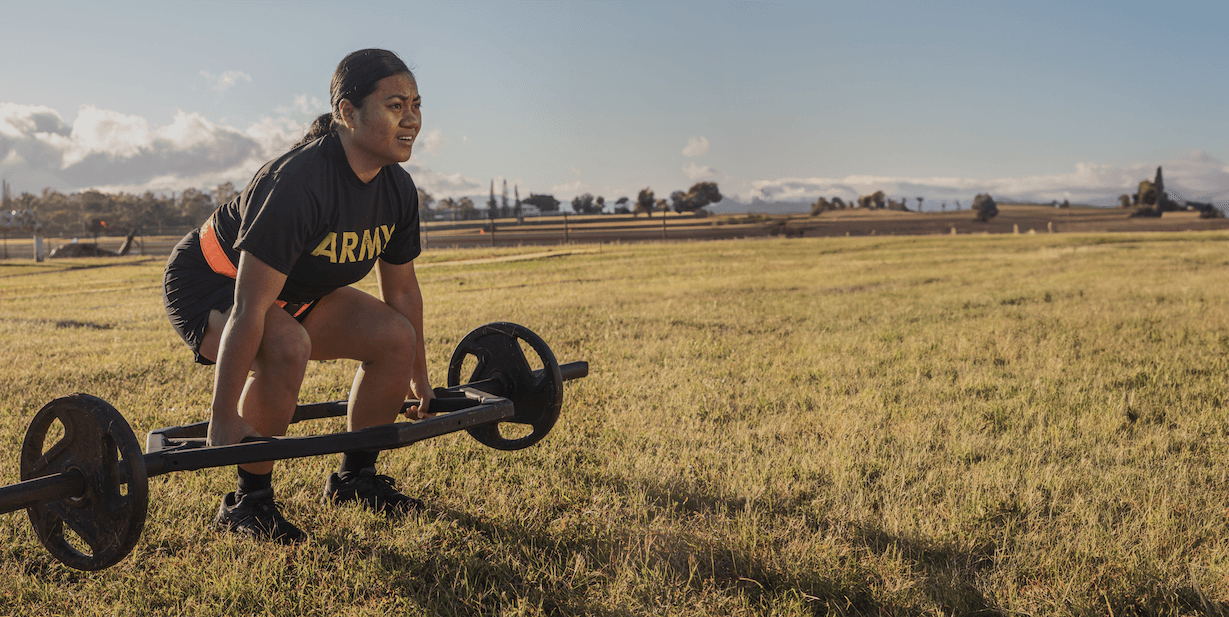 A determined female soldier prepares for a deadlift during an ACFT event, with weights set against a grassy field backdrop.