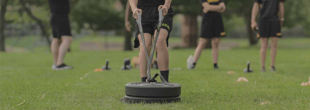 Soldier in 'Sprint-Drag-Carry' ACFT event, pulling weight on grass field, with other participants in the background.