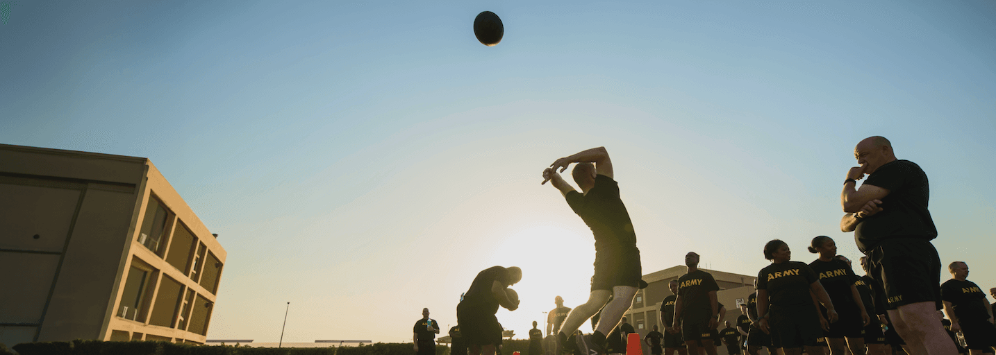 Soldier performs standing power throw event in Army Combat Fitness Test (ACFT) at sunset, with other participants in the background.